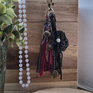May include: A decorative keychain with a black lace flower, pearl accents, and dangling bronze-colored charms. The keychain features burgundy and black ribbons and is attached to a wooden background. A strand of white beads hangs nearby.