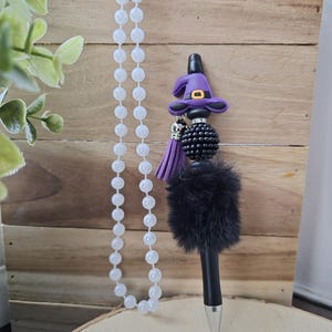 Könnte beinhalten: Ein schwarzer Stift mit einem Hexenhut-Anhänger, schwarzem Fell und einer lila Quaste. Der Stift hat eine silberne Spitze und einen schwarzen Schaft. Eine weiße Perlenkette hängt im Hintergrund. Der Stift ist mit einem lila Hexenhut mit goldener Schnalle verziert.
