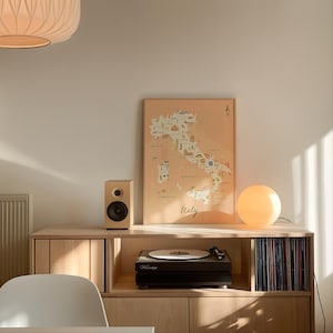 May include: A framed map of Italy with a light peach background, featuring illustrated landmarks and the word Italy in script. The map sits on a light wood media console next to a wooden speaker and a round white lamp, above a black record player.