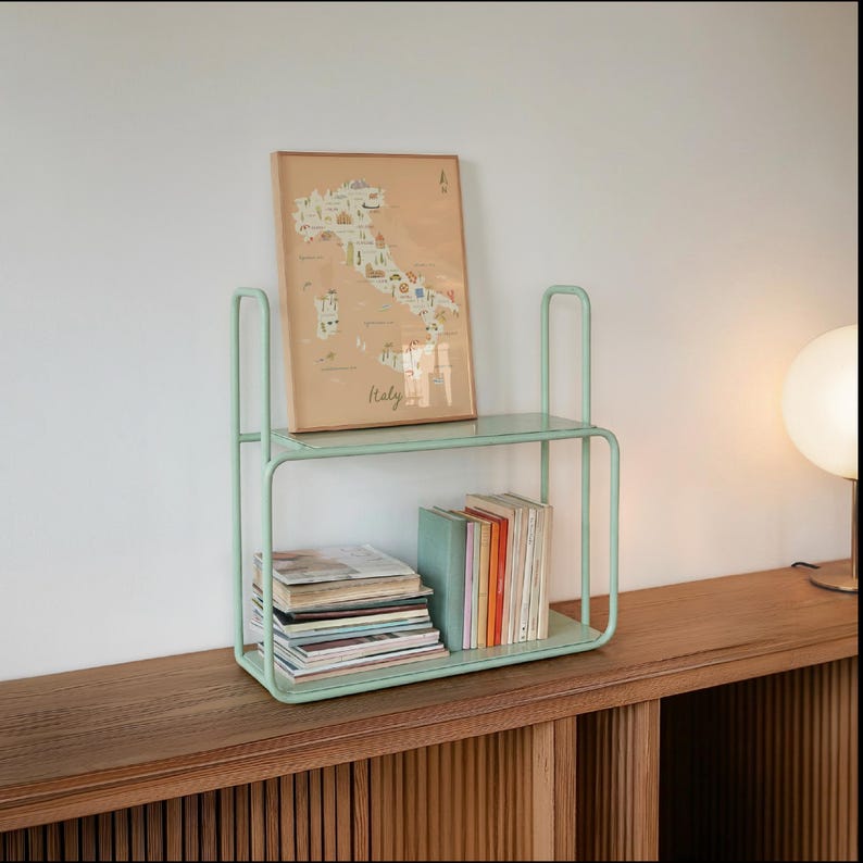 May include: A light mint green metal two-tier shelf with rounded edges. The top shelf holds a framed map print labeled Italy, while the bottom shelf contains a stack of magazines and a row of books with various colored spines arranged vertically.