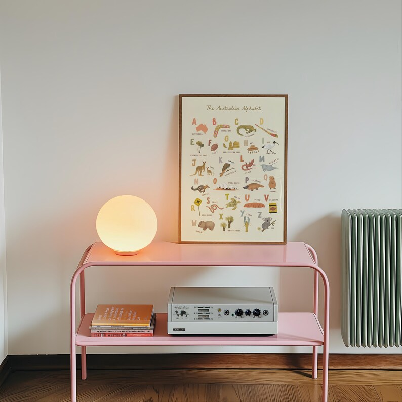 Puede incluir: Una mesa consola de metal rosa con dos estantes, que sostiene una l&aacute;mpara esf&eacute;rica brillante, una pila de libros y un dispositivo electr&oacute;nico rectangular. Un p&oacute;ster enmarcado titulado The Australian Alphabet con ilustraciones est&aacute; colocado encima.