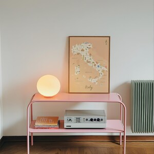 May include: A pink metal two-tier console table holding a glowing spherical lamp, a framed map of Italy with illustrations, a stack of books, and a grey electronic device with knobs and switches. The map features the text Italy in script.