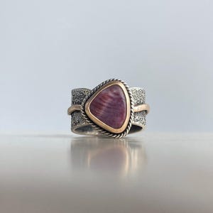 May include: A silver ring with a triangular, purple gemstone set in a gold bezel. The band has a textured design with gold accents. The ring is displayed on a reflective surface.
