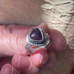 May include: A silver ring featuring a triangular, deep purple cabochon stone set in a bezel. The metal band displays etched decorative patterns along the sides and around the stone setting, creating a textured appearance on the silver surface.