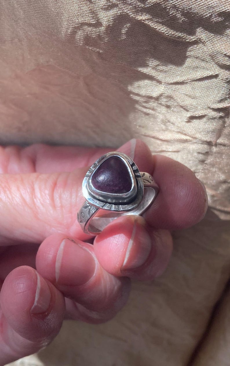 May include: A silver ring featuring a triangular, deep purple cabochon stone set in a bezel. The metal band displays etched decorative patterns along the sides and around the stone setting, creating a textured appearance on the silver surface.