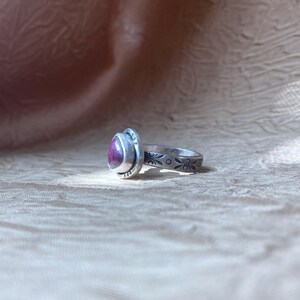 May include: A silver ring featuring a teardrop-shaped magenta stone set in a bezel. The band is decorated with a repeating pattern of small etched floral motifs and dots, showing a dark oxidized finish within the engraved details of the metal.