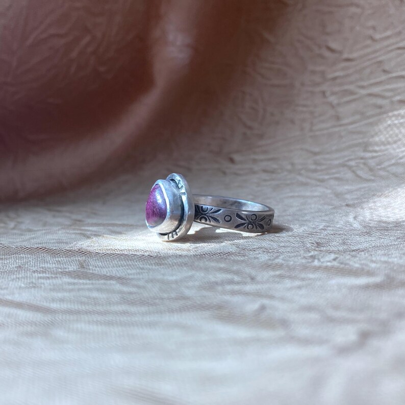 May include: A silver ring featuring a teardrop-shaped magenta stone set in a bezel. The band is decorated with a repeating pattern of small etched floral motifs and dots, showing a dark oxidized finish within the engraved details of the metal.