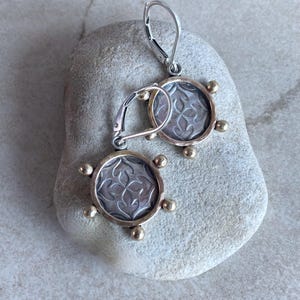 May include: A pair of silver and gold-toned earrings. Each earring features a circular design with a floral pattern in the center, surrounded by a gold-toned border and small gold accents. The earrings have a lever-back closure.