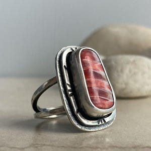 May include: A silver ring with an oval, red and white stone. The stone is set in a silver bezel with a decorative border. The ring has a split shank design. The ring is on a light-colored surface with blurred stones in the background.