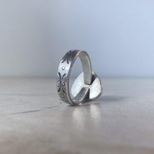 May include: A silver-toned metal ring featuring a band with an engraved pattern of small floral motifs and circular dots. The ring has a flat, heart-shaped element attached to the side of the band, displaying a smooth, reflective surface finish.