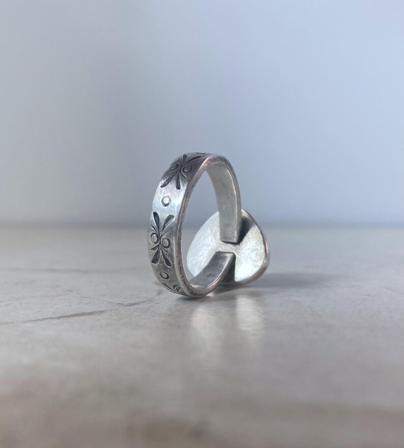 May include: A silver-toned metal ring featuring a band with an engraved pattern of small floral motifs and circular dots. The ring has a flat, heart-shaped element attached to the side of the band, displaying a smooth, reflective surface finish.