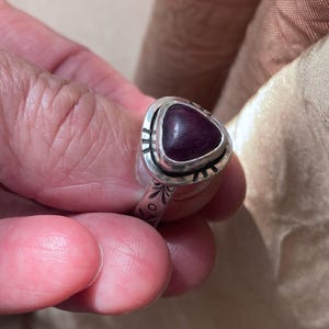May include: A silver ring featuring a triangular, deep purple cabochon stone set in a bezel. The metal band and the setting surrounding the stone are decorated with etched, dark-colored linear and circular patterns along the surface.