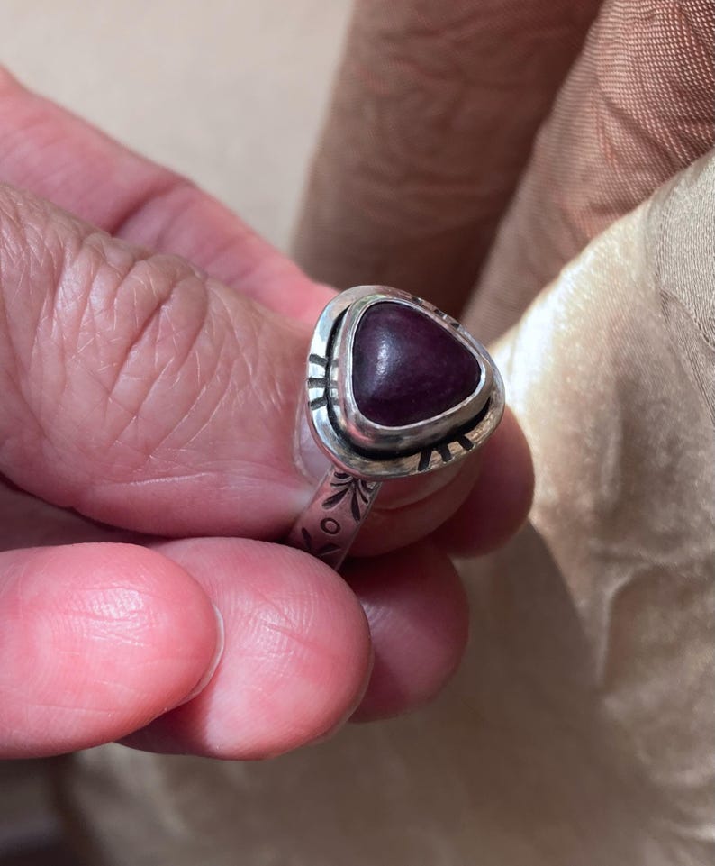 May include: A silver ring featuring a triangular, deep purple cabochon stone set in a bezel. The metal band and the setting surrounding the stone are decorated with etched, dark-colored linear and circular patterns along the surface.