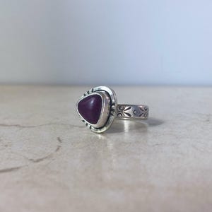 May include: A silver ring featuring a triangular, deep purple stone set in a smooth bezel. The metal band is decorated with etched floral patterns and small lines around the stone setting, creating a detailed, textured appearance on the silver.