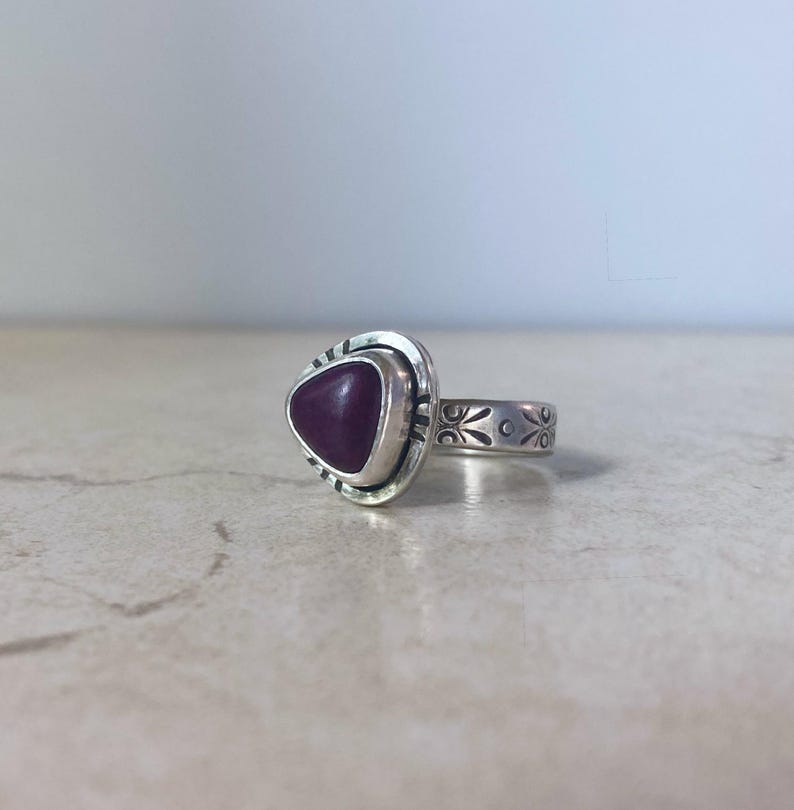 May include: A silver ring featuring a triangular, deep purple stone set in a smooth bezel. The metal band is decorated with etched floral patterns and small lines around the stone setting, creating a detailed, textured appearance on the silver.