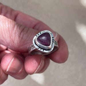 May include: A silver ring featuring a triangular, deep magenta-colored stone set in a bezel. The silver band has a textured, stamped pattern around the setting and along the sides of the band, creating a detailed, metallic finish on the ring.