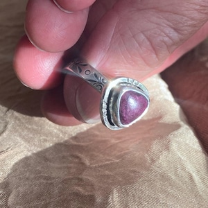 May include: A silver ring featuring a triangular, deep magenta-colored stone set in a bezel. The band of the ring is decorated with engraved floral patterns and small circular accents, showcasing a polished metallic finish throughout the piece.