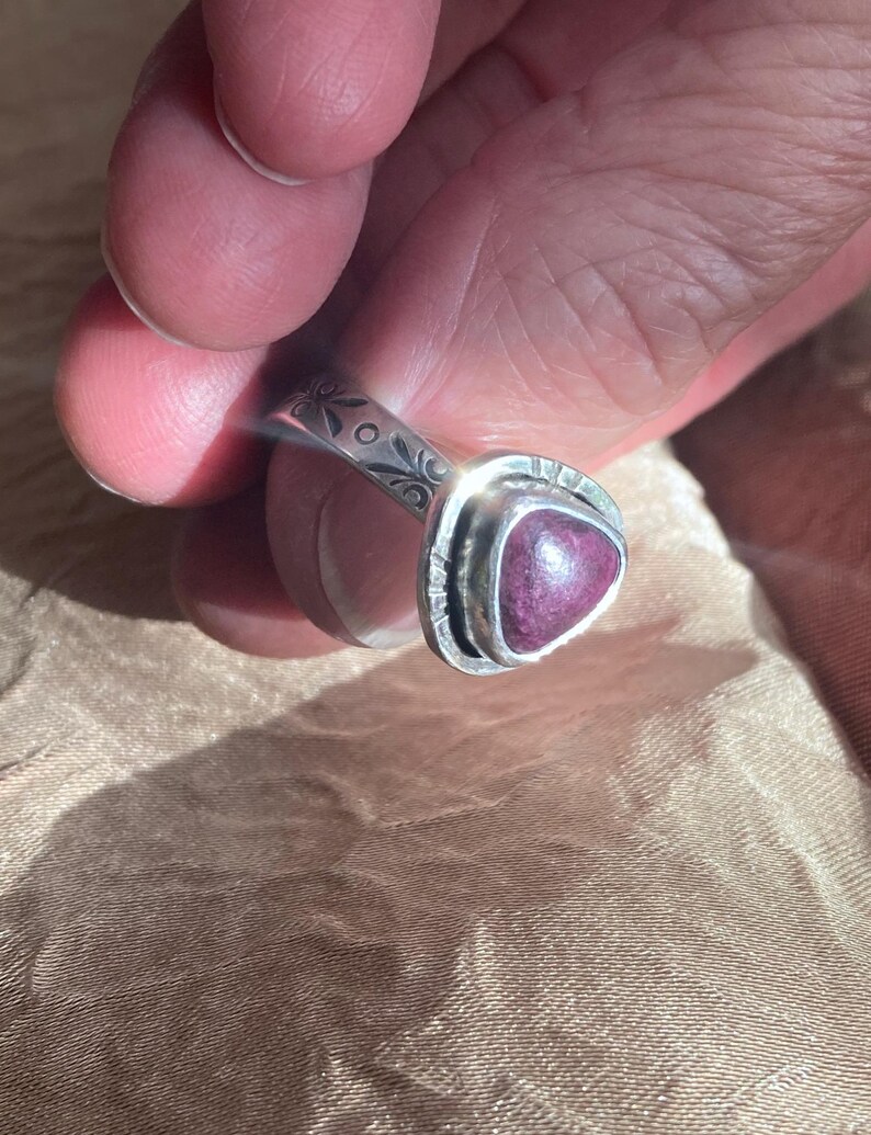 May include: A silver ring featuring a triangular, deep magenta-colored stone set in a bezel. The band of the ring is decorated with engraved floral patterns and small circular accents, showcasing a polished metallic finish throughout the piece.