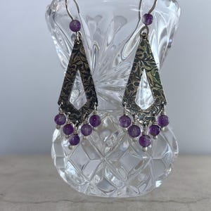 May include: Silver-toned dangle earrings with a textured, triangular design. Each earring features a series of small, round, purple beads hanging from the bottom. The earrings are displayed in a clear, cut-glass vase.