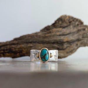 May include: A silver ring with a turquoise stone set in a gold bezel. The ring has a textured band with etched details. The background features a piece of driftwood against a white backdrop.