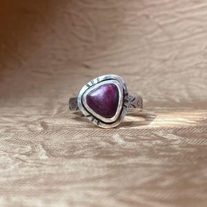 May include: A silver ring featuring a triangular, deep magenta-colored stone set in a bezel. The metal band displays etched markings around the stone setting and along the sides of the band, creating a textured appearance on the silver surface.