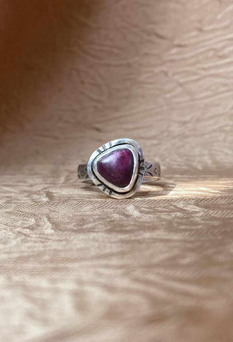 May include: A silver ring featuring a triangular, deep magenta-colored stone set in a bezel. The metal band displays etched markings around the stone setting and along the sides of the band, creating a textured appearance on the silver surface.