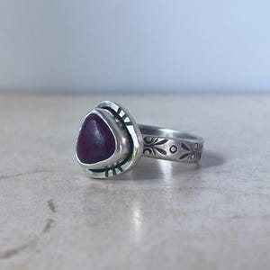 May include: A silver ring featuring a triangular, deep red stone set in a bezel. The silver band is decorated with etched, repeating floral-like patterns and small vertical lines around the stone setting, creating a textured, oxidized appearance.