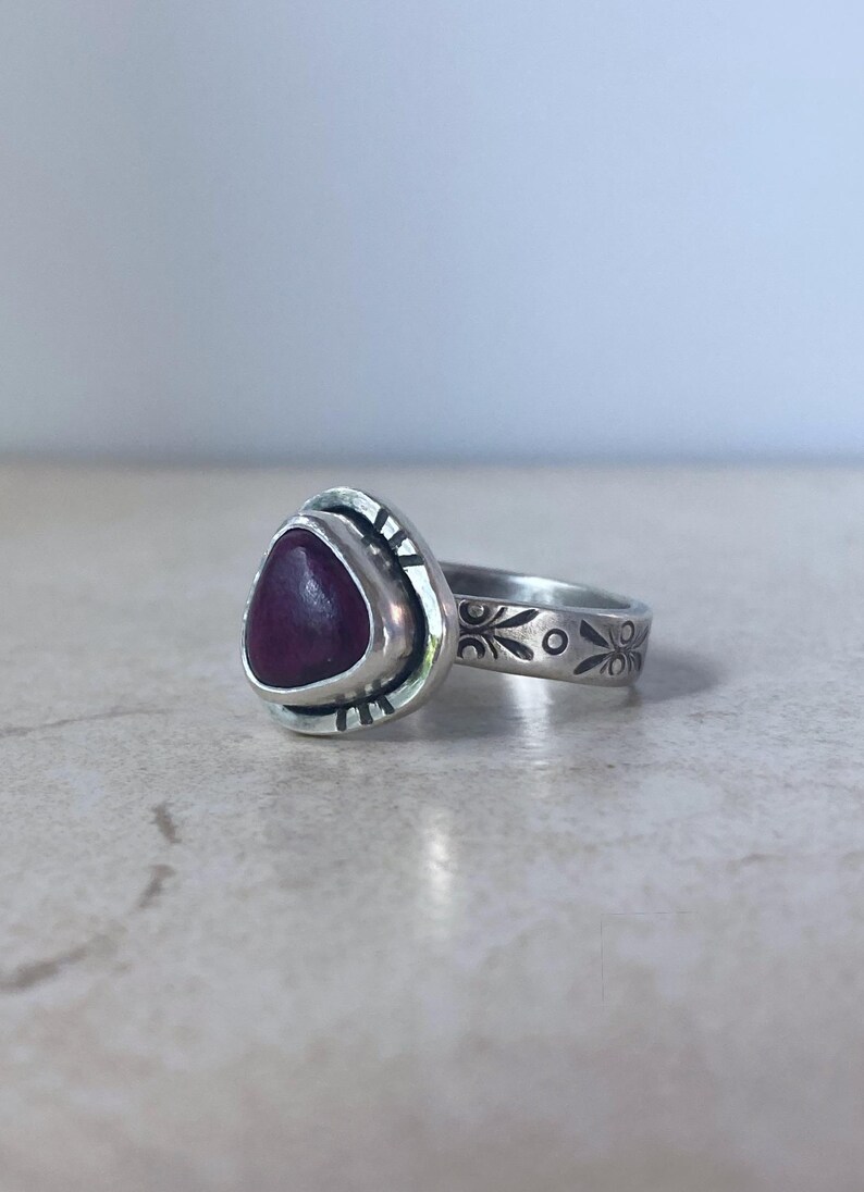 May include: A silver ring featuring a triangular, deep red stone set in a bezel. The silver band is decorated with etched, repeating floral-like patterns and small vertical lines around the stone setting, creating a textured, oxidized appearance.