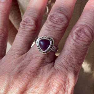 May include: A silver ring featuring a triangular, deep magenta-colored stone set in a bezel. The silver band has a polished finish with small, etched decorative markings around the stone setting and along the sides of the band.