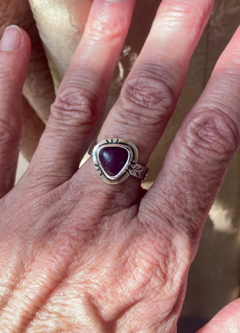May include: A silver ring featuring a triangular, deep magenta-colored stone set in a bezel. The silver band has a polished finish with small, etched decorative markings around the stone setting and along the sides of the band.
