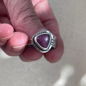 May include: A silver ring featuring a triangular, deep magenta-colored stone set in a bezel. The silver band has a textured, stamped pattern along its outer surface, surrounding the central stone with a polished, metallic finish and distinct edges.