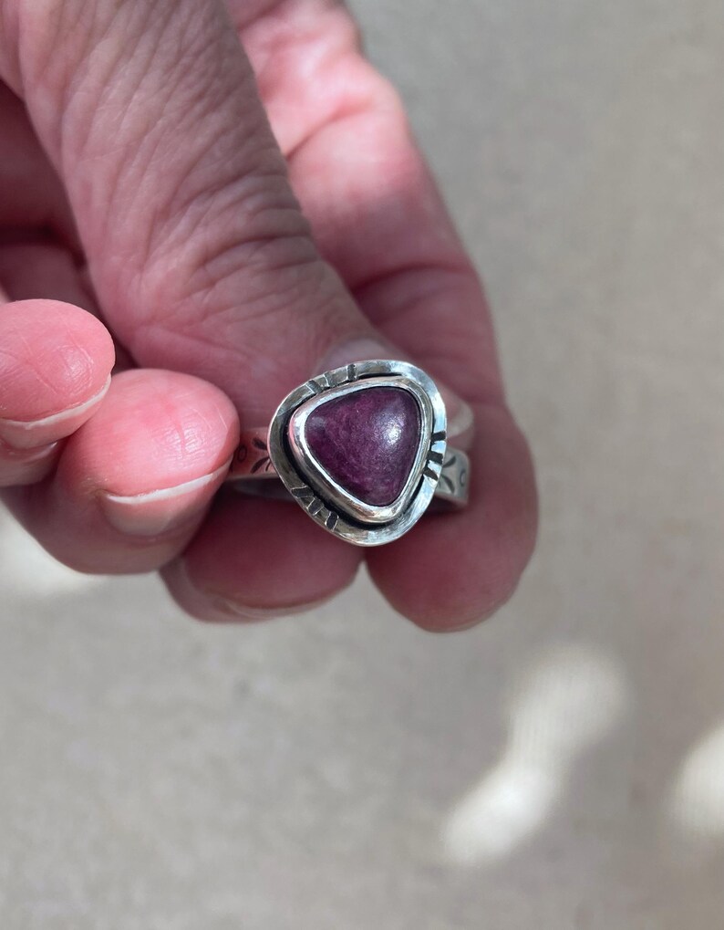 May include: A silver ring featuring a triangular, deep magenta-colored stone set in a bezel. The silver band has a textured, stamped pattern along its outer surface, surrounding the central stone with a polished, metallic finish and distinct edges.