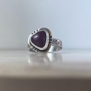 May include: A silver ring featuring a triangular, deep purple cabochon stone set in a bezel. The metal band displays engraved floral patterns and decorative notches around the stone setting, creating a textured appearance on the silver surface.