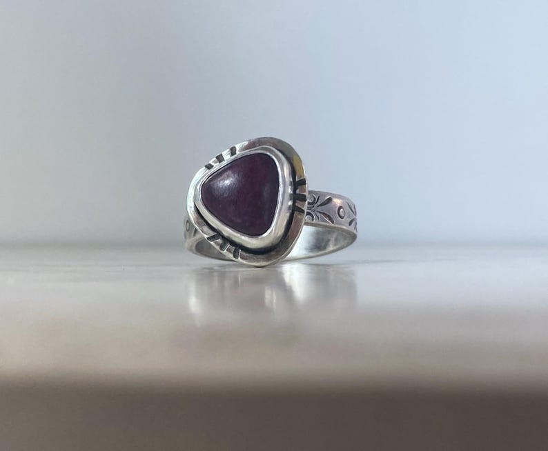 May include: A silver ring featuring a triangular, deep purple cabochon stone set in a bezel. The metal band displays engraved floral patterns and decorative notches around the stone setting, creating a textured appearance on the silver surface.