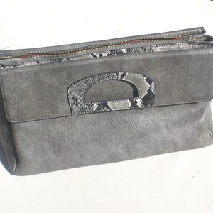Könnte beinhalten: Eine graue Clutch mit einem Schlangenleder-Mustergriff. Die Clutch hat einen Reißverschluss und einen langen Riemen.