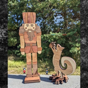 May include: A wooden nutcracker figurine with a red and brown hat, a brown beard, and a brown and gold uniform. A wooden squirrel figurine with a brown tail and a brown body is holding a nut in its paws. Both figurines are standing on a wooden base.