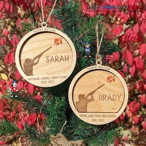 May include: Two wooden Christmas ornaments with a silhouette of a person holding a shotgun and a clay pigeon target. The top ornament says "Nothin' But Dust" and "SARAH" with "SOUTHLAKE CARROLL TARGET TEAM 2022-2023". The bottom ornament says "Nothin' But Dust" and "BRADY" with "SPRING LAKE PARK HIGH SCHOOL 2018-2022".