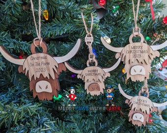 Puede incluir: Cuatro adornos de madera de vaca de las tierras altas con diferentes saludos navideños. Los adornos son marrones y tienen una cara marrón claro. Los adornos están colgados de un árbol de Navidad.