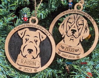 Puede incluir: Dos adornos de perro de madera. El primer adorno es marrón y negro con la cabeza de un perro y el texto "Newest Member of the Pack" y "RAISIN 2023". El segundo adorno es marrón con la cabeza de un perro y el texto "In Loving Memory Of ZEUS 2007-2018".