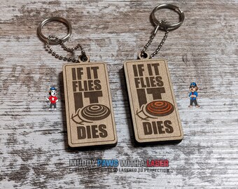 Puede incluir: Dos llaveros de madera con fondo marrón y el texto "If it flies it dies" y un gráfico de una paloma de arcilla. Los llaveros están unidos a cadenas plateadas con un anillo de llave plateado.
