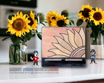 Puede incluir: Porta llaves de madera con un diseño de girasol cortado con láser. El porta llaves tiene dos ranuras para llaves y está pintado en tonos marrones y negros. El texto "Muddy Paws with a Laser" está impreso debajo del porta llaves.
