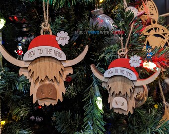 Puede incluir: Dos adornos navideños de madera con forma de cabeza de vaca Highland, que llevan un gorro de Papá Noel rojo con el texto "New to the Herd" y el año "2023".