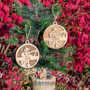 May include: Two wooden Christmas ornaments with a shooting sports theme. Each ornament features a silhouette of a person aiming a gun, the name of a person, and the year 2024. The ornaments are hanging on a small evergreen tree with a red and pink background.