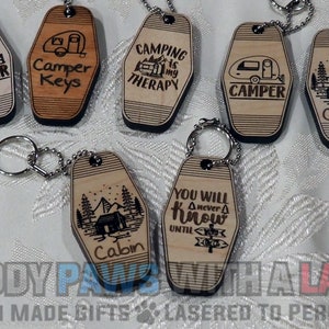 May include: A set of six wooden keychains with various camping-themed designs. The keychains are shaped like motel keys and feature laser-engraved images and text, including "Camper", "Cabin", "Camping is my therapy", and "You will never know until you go".