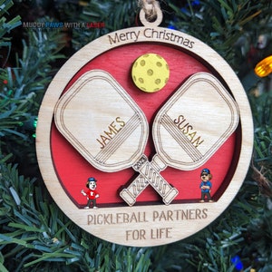 May include: A wooden Christmas ornament with a red background and two crossed pickleball paddles. The paddles are labeled "JAMES" and "SUSAN". The ornament also features a small pickleball, the year "2023", and the text "Pickleball Partners for Life".