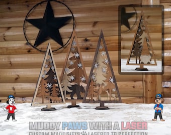 Puede incluir: Decoraciones de árboles de Navidad de madera con un diseño en capas. Cada árbol presenta una escena forestal diferente con animales y copos de nieve. Los árboles son marrones y marrón claro, con una base marrón oscuro. El texto "MUDDY PAWS WITH A LASER" está en la parte inferior.