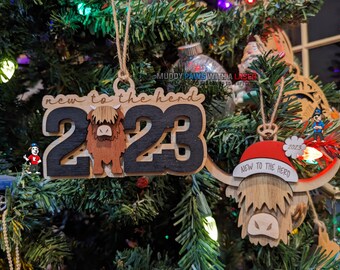 Puede incluir: Dos adornos navideños de madera con diseño de vaca Highland. El primer adorno es negro y marrón con los números "2023" y el texto "new to the herd". El segundo adorno es una cabeza de vaca Highland marrón con un gorro de Papá Noel rojo con el texto "new to the herd" y el número "2023".