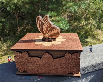 Puede incluir: Caja de madera marrón con una mariposa cortada con láser en la parte superior. La caja tiene diseños florales en la parte delantera y superior. La mariposa está hecha de madera en capas en tonos marrones y beige. El texto "MUDDY PAWS WITH A LASER" está en la parte inferior.