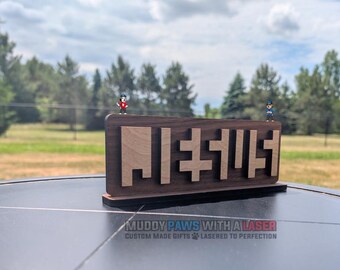 Puede incluir: Un letrero rectangular de madera con una base de color marrón oscuro y letras en relieve de color marrón claro que forman la palabra JESUS. Dos pequeñas figuras de perros de dibujos animados están sobre el borde superior del letrero.