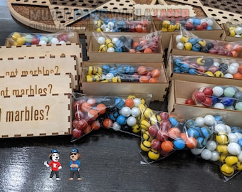 Puede incluir: Canicas de colores en cajas y bolsas de madera. Las canicas son de varios colores, incluyendo rojo, azul, amarillo y blanco. Las cajas tienen un diseño cortado con láser y el texto "...not marbles? ...not marbles? ...not marbles?" es visible.